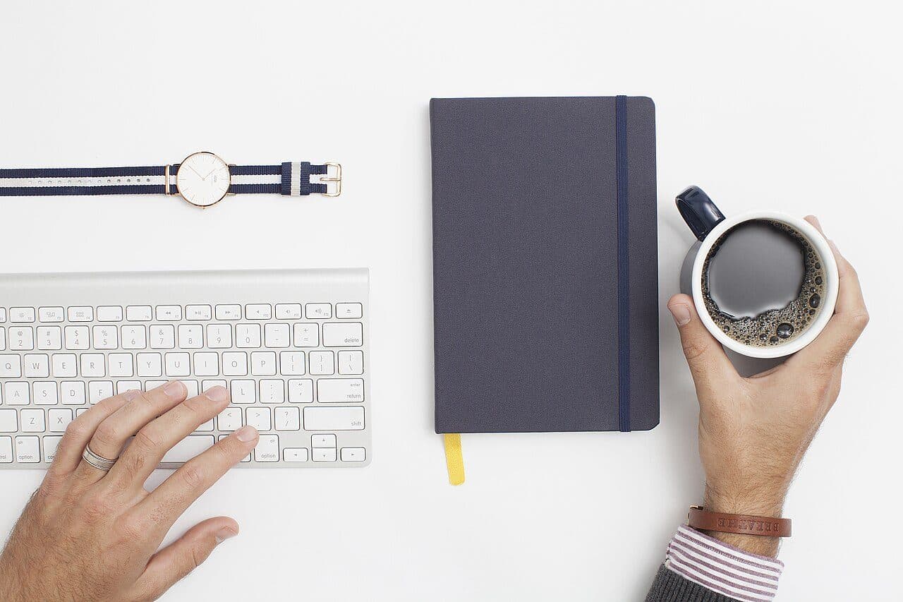 Vue de dessus d'un bureau : clavier, carnet, montre et tasse de café tenue par une main