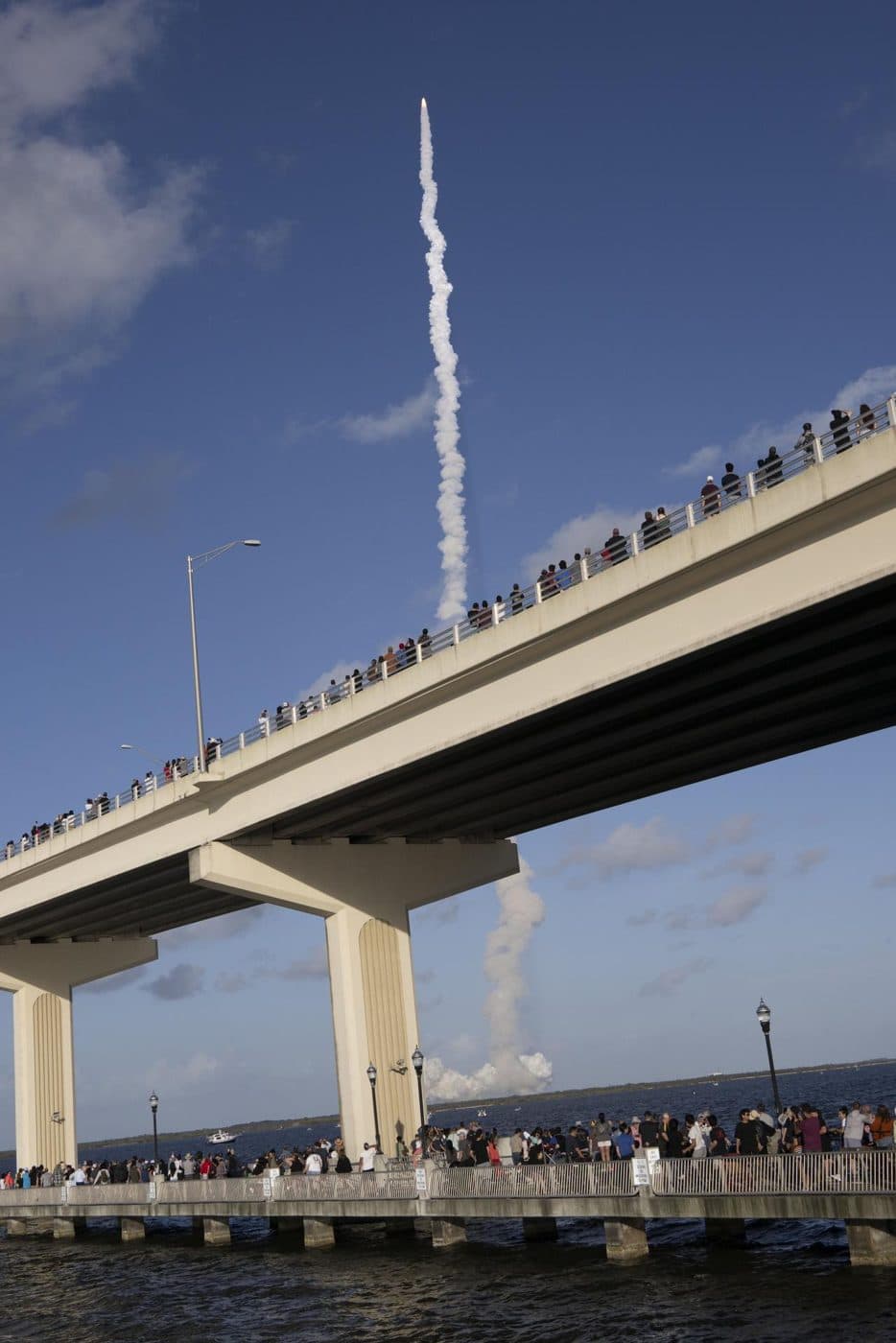 Foule massée sur un pont et une jetée, sous le panache d'une fusée au décollage